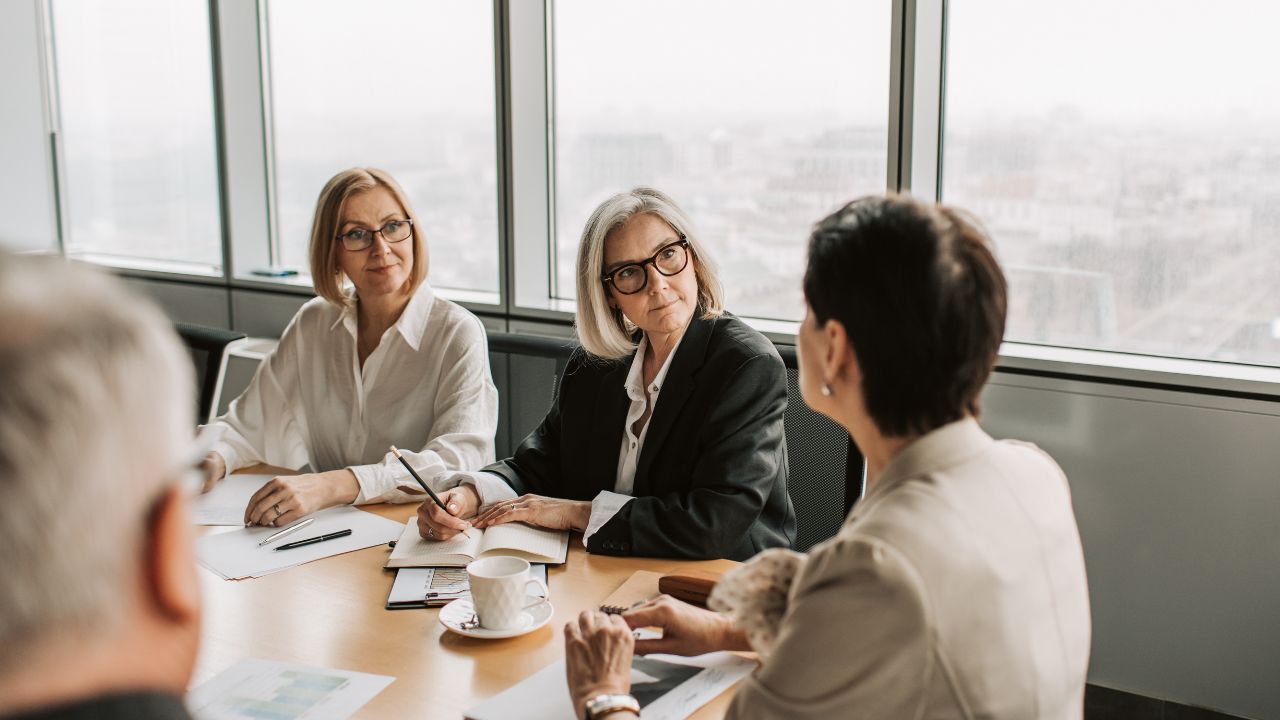 Four adults sit at a conference table in a meeting room with large windows, dressed professionally in attire suitable for what to wear to a board meeting, listening and taking notes during a discussion.
